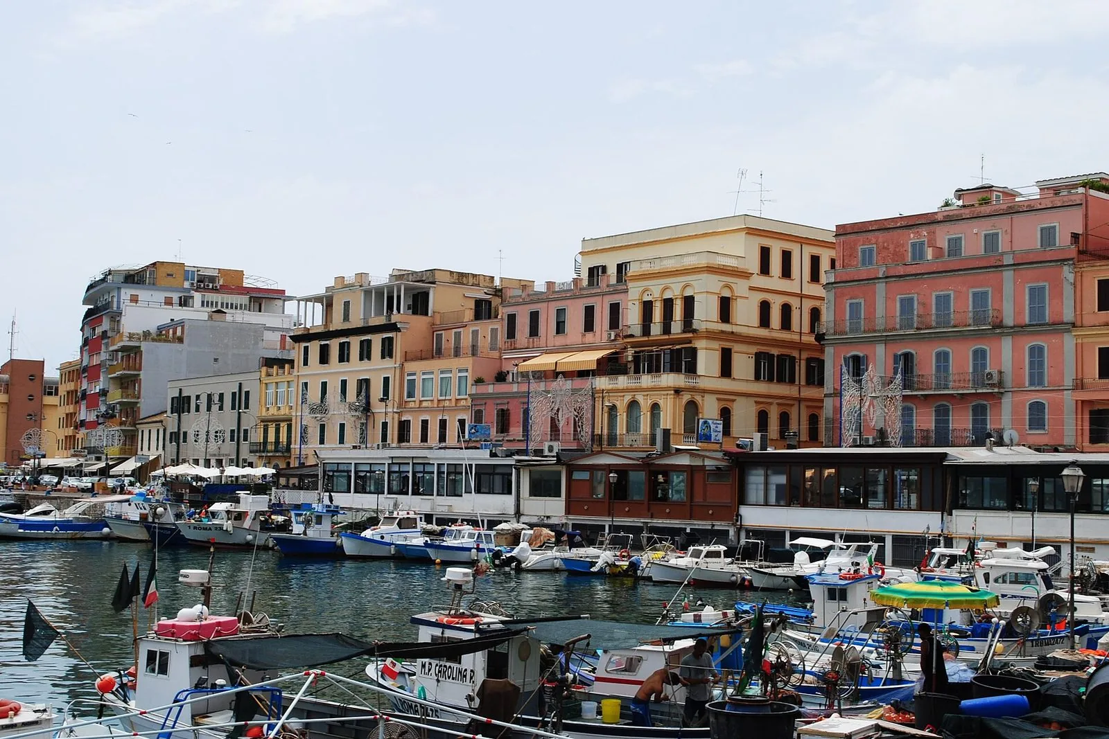 Il porto di Anzio con le barche da pesca ormeggiate in primo piano, reti e attrezzature sul ponte, e alle spalle la fila di palazzi color pastello affacciati sulla banchina