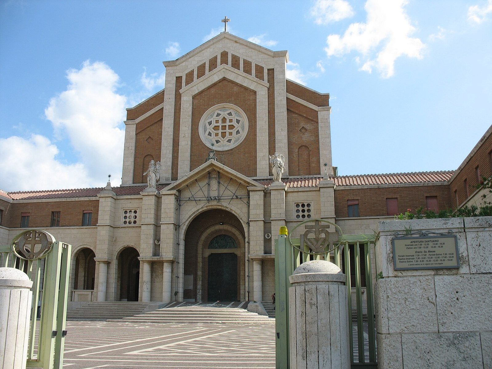 La facciata del santuario di Nostra Signora delle Grazie e Santa Maria Goretti a Nettuno, in mattoni e pietra chiara con il rosone centrale e due statue di angeli, vista dal cancello d'ingresso sotto un cielo azzurro con nuvole