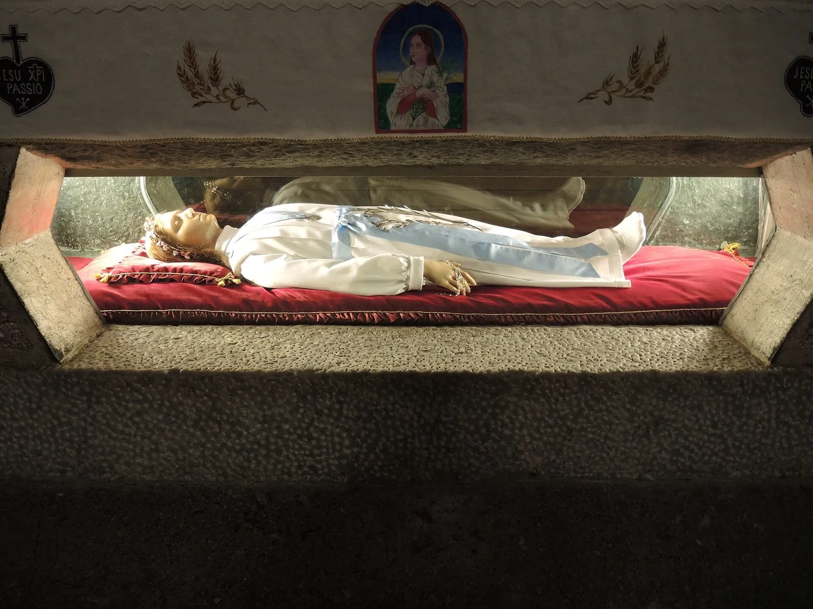 L'urna di Santa Maria Goretti nella cripta del santuario di Nettuno: una figura distesa in abito bianco con fascia azzurra su un cuscino rosso, dietro il vetro, sotto un drappo ricamato con l'immagine della santa e spighe di grano